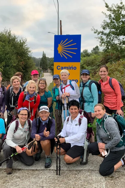 A group of fifteen hikers with backpacks and trekking poles posed on a roadside near a "Camiño" sign, surrounded by greenery.