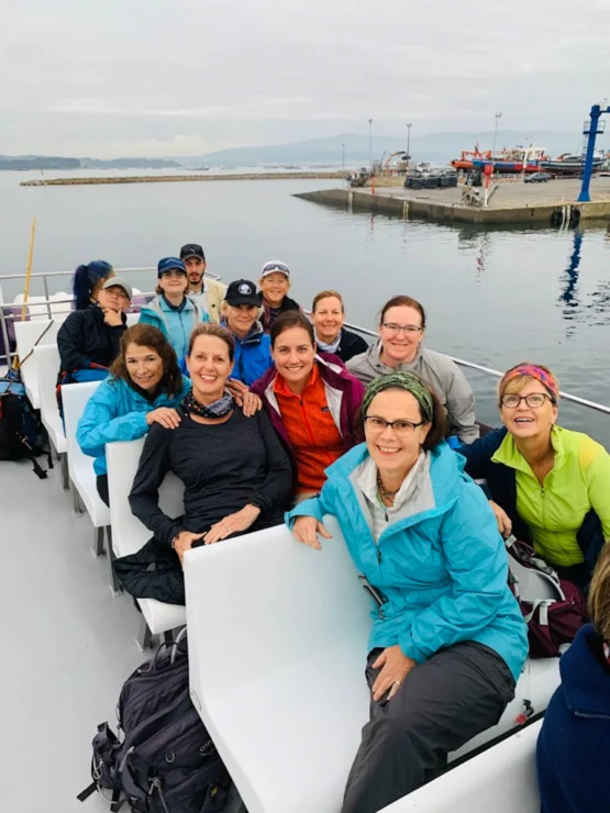 A group of people, mostly women, sit on a boat with harbor and docks in the background. They are dressed in outdoor jackets and appear to be enjoying their trip.