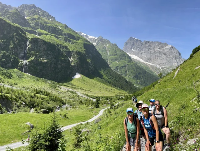 A group of hikers poses on a green mountain trail with a waterfall and rocky peaks in the background under a clear blue sky.