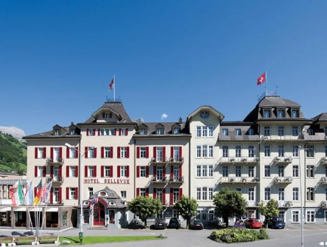 A large hotel with a traditional design, featuring red shutters and several balconies. It has Swiss flags on the roof and is set against a backdrop of mountains and clear blue sky.