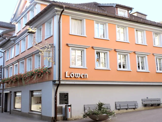 Three-story building with an orange facade, labeled "Löwen." White-framed windows, floral decorations, and a hanging sign. A street and benches are in front.