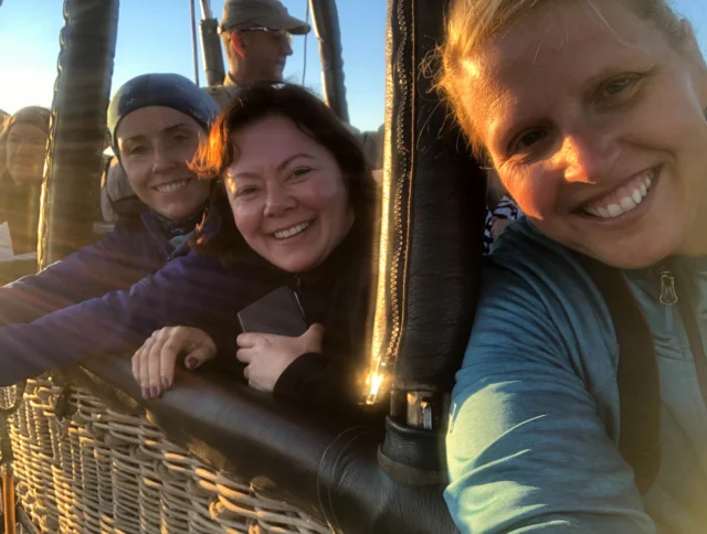 A group of people smile while standing in a hot air balloon basket during a sunny day.