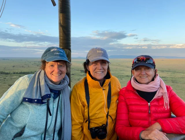 Three people in jackets and hats smile while standing in a hot air balloon basket, with a vast landscape and a blue sky in the background.