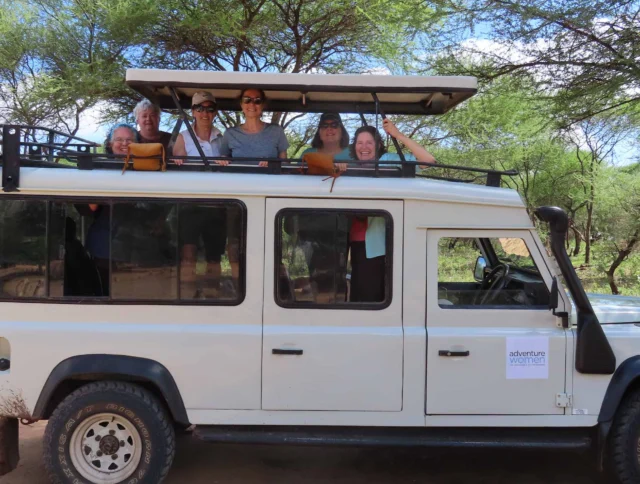 Group of people inside a white safari vehicle with an open roof, parked near green trees.