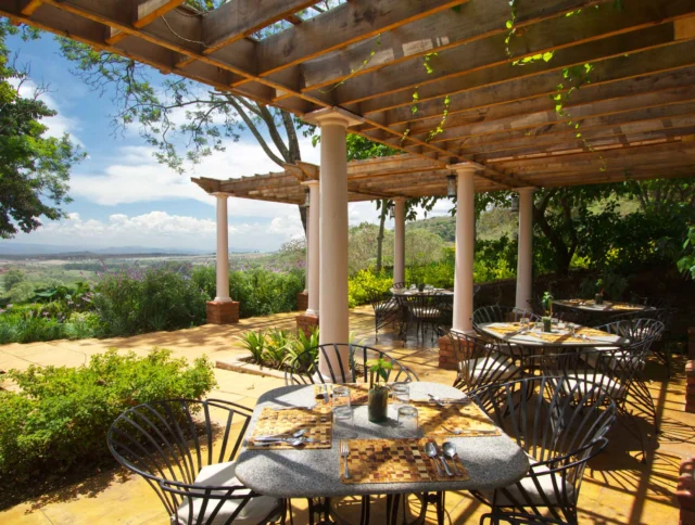 Outdoor restaurant patio with round tables set for four under a wooden pergola, surrounded by lush greenery and a scenic view of mountains in the background.