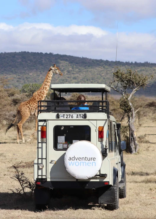 A safari vehicle with "Adventure Women" on the spare tire cover parks in a savannah as a giraffe walks by.