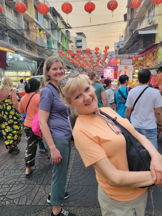 Two people pose on a bustling street decorated with red lanterns. Others walk around them, and buildings line the street.