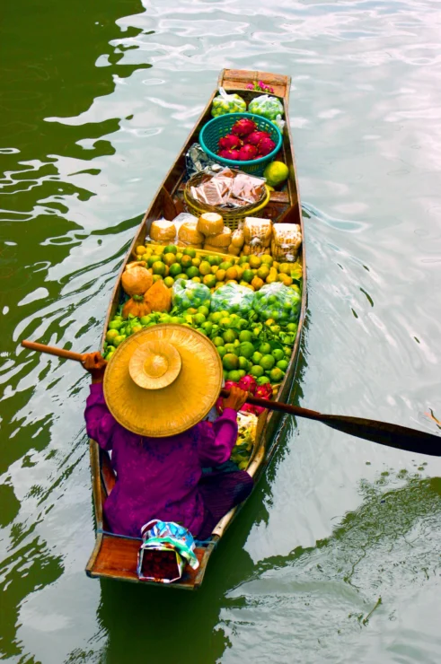 A person in a wide-brimmed hat rows a wooden boat filled with various fruits and goods on a body of water.