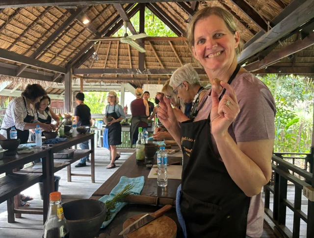 A smiling woman in an apron stands in an open-air kitchen. Others are cooking at wooden tables in the background.