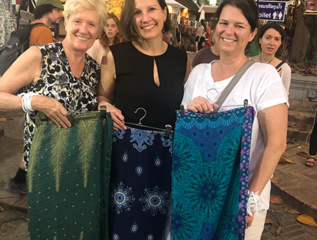 Three women smiling and holding colorful fabrics at an outdoor market.