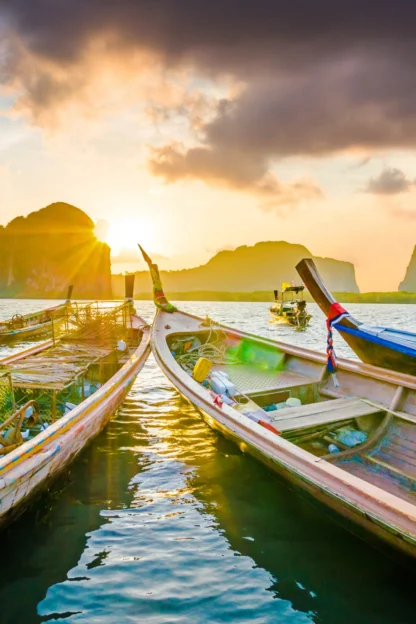 Several long-tail boats on water with dramatic sky and rocky islands in the background at sunset.