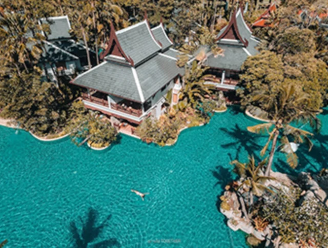 Aerial view of a person swimming in a large, tropical resort pool surrounded by lush greenery and traditional-style buildings.