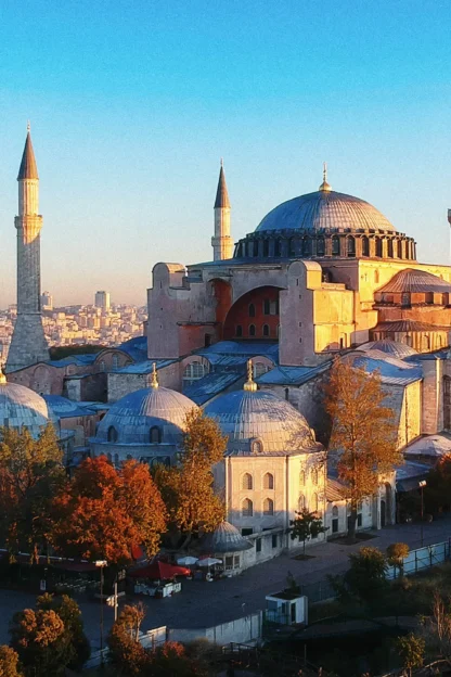 Aerial view of the Hagia Sophia in Istanbul, showing its large dome, four minarets, and surrounding trees with a cityscape backdrop under a clear sky.