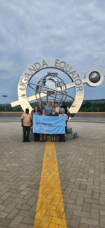 Group of people standing at the Uganda Equator marker, holding a banner that reads "Adventure Voyaging," with a paved area and cloudy sky in the background.