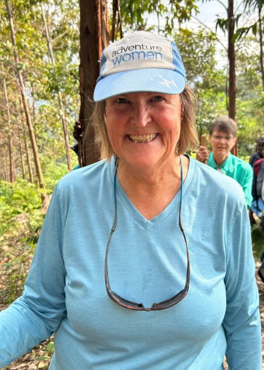 Woman wearing a blue "adventure women" cap and sunglasses around her neck smiles outdoors, with trees and another person in the background.