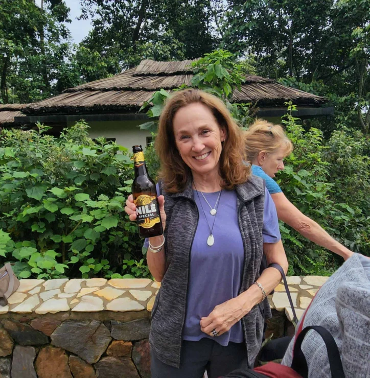 A woman smiles while holding a bottle of Nile Special beer in front of lush greenery and a stone wall. Another person is partially visible in the background.