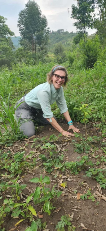 Person kneeling in a green field, planting or tending to a young plant, surrounded by vegetation and hills in the background.