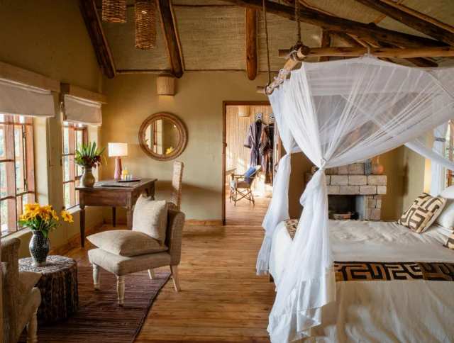 Cozy bedroom with a canopy bed, wooden flooring, and rustic decor. Includes a writing desk, chairs, vase with yellow flowers, and a round mirror. Large windows provide natural light.