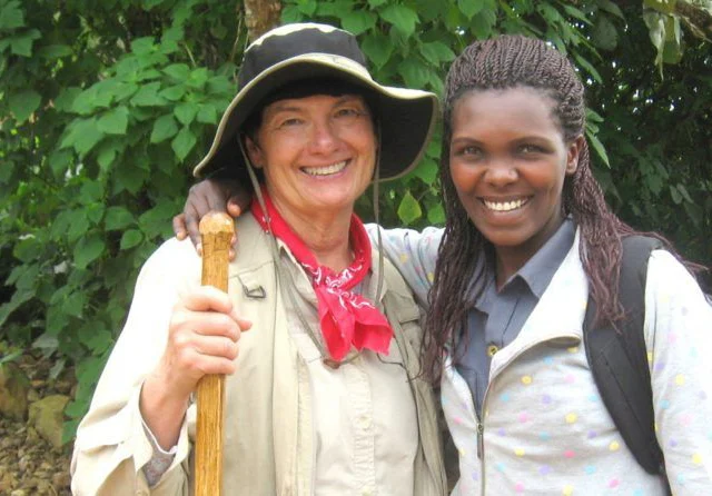 Two women smiling, one wearing a hat and holding a walking stick, standing outdoors in front of greenery.