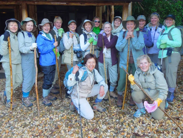 A group of hikers pose together with walking sticks, wearing outdoor gear and hats, standing on a gravel path in front of a wooden building.