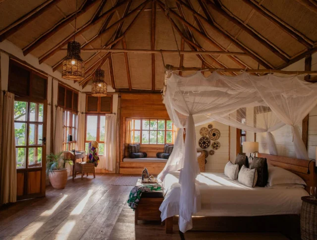 A spacious bedroom with a canopy bed, wooden floors, and large windows. A seating area and decor elements are visible, with natural light streaming in.