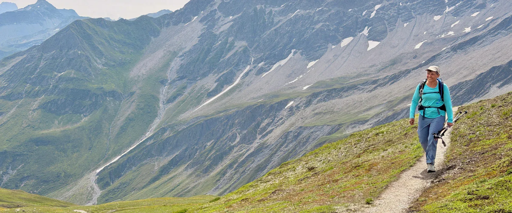 A person with a backpack walks along a mountain trail on a grassy slope, with rocky, snow-patched mountains in the background.