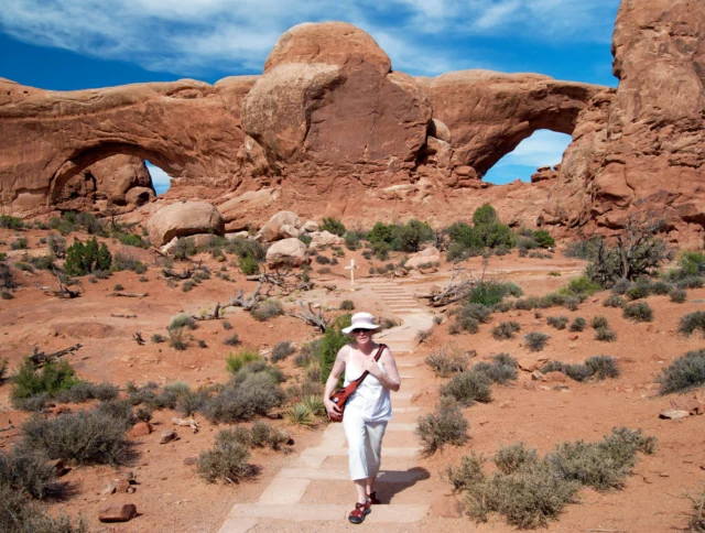 A person in white clothing and a sunhat walks on a stone path in a desert landscape with large rock formations and a bright blue sky.