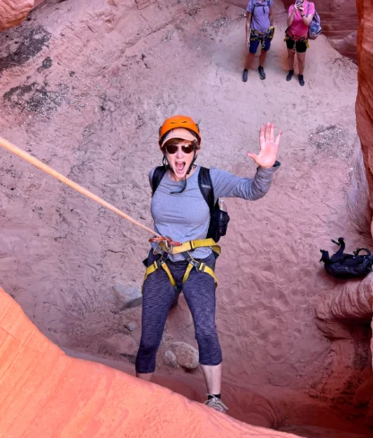 Person in climbing gear waves while rappelling down a rocky canyon, with two people in similar gear standing at the top.