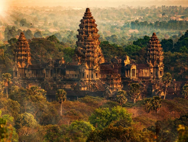 Angkor Wat temple complex surrounded by lush greenery at sunrise, with intricate towers and ancient stone architecture visible.