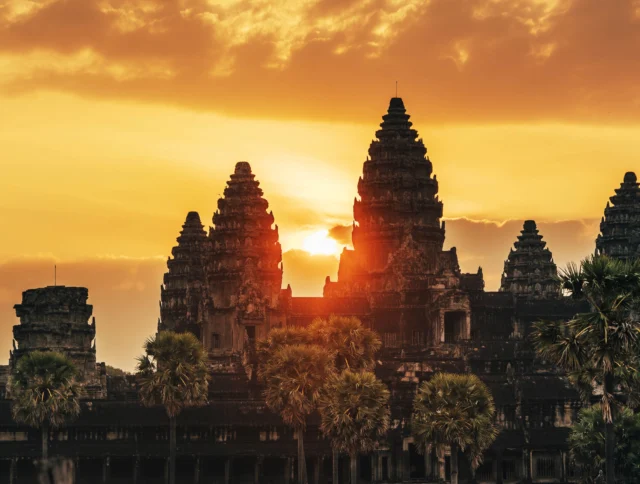 Sunset over Angkor Wat, Cambodia, with an orange sky and silhouetted temple spires surrounded by palm trees.