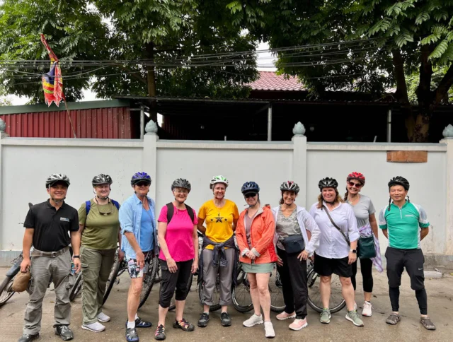 A group of people wearing helmets and casual clothing pose together in front of a white wall and trees. Some hold cycling gear.