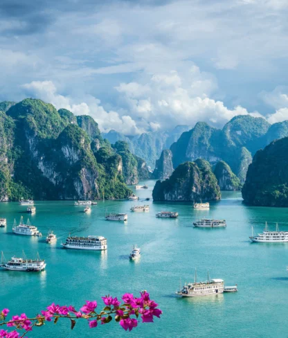 Boats navigate between limestone islands under a cloudy sky in a turquoise bay, with pink flowers in the foreground.