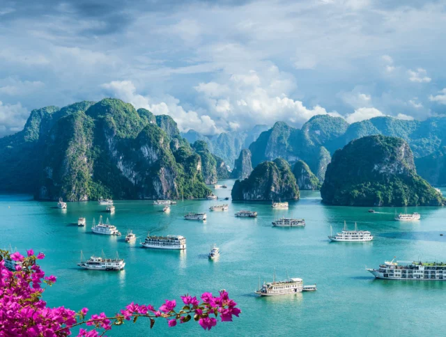 Boats navigate between limestone islands under a cloudy sky in a turquoise bay, with pink flowers in the foreground.