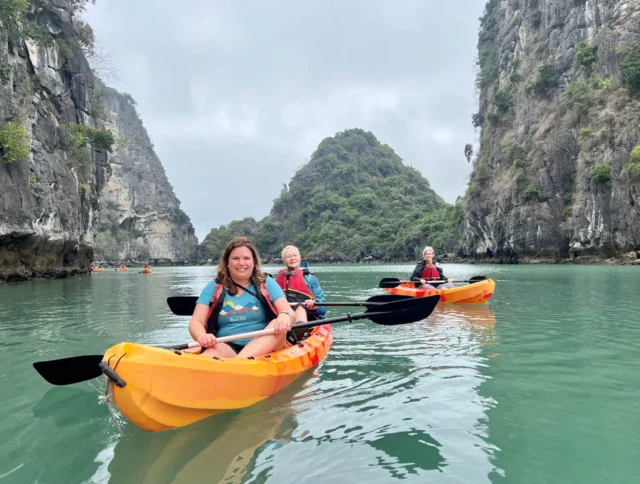 Three people kayaking on calm water between rocky cliffs and lush hills under a cloudy sky.