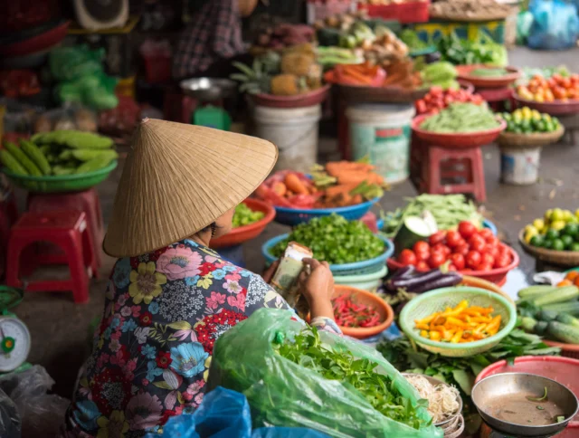 A person in a conical hat sits at a vegetable market stall, surrounded by various fresh produce, including tomatoes, peppers, and greens.