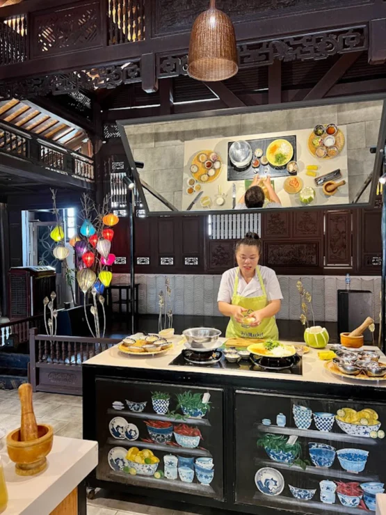 A person in a green apron prepares food in a kitchen with wooden decor, colorful lanterns, and various dishes on display.