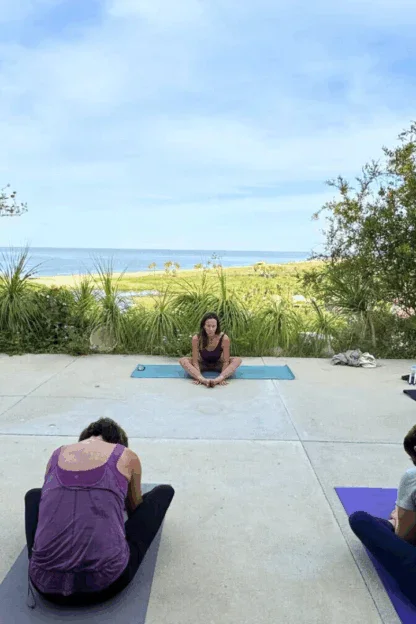 A yoga instructor leads a group of six people in seated poses on yoga mats outdoors, with greenery and a view of the ocean in the background.