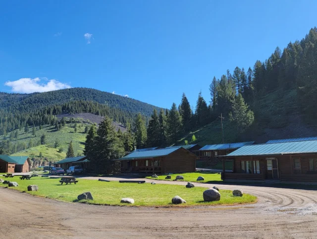 Rustic cabins with green roofs are nestled in a lush, forested valley under a clear blue sky. A dirt road curves in front, with picnic tables and scattered rocks on grassy areas.