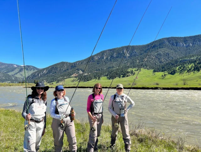 Four people in fishing gear stand by a river, holding fishing rods. Hills and a clear blue sky are in the background.