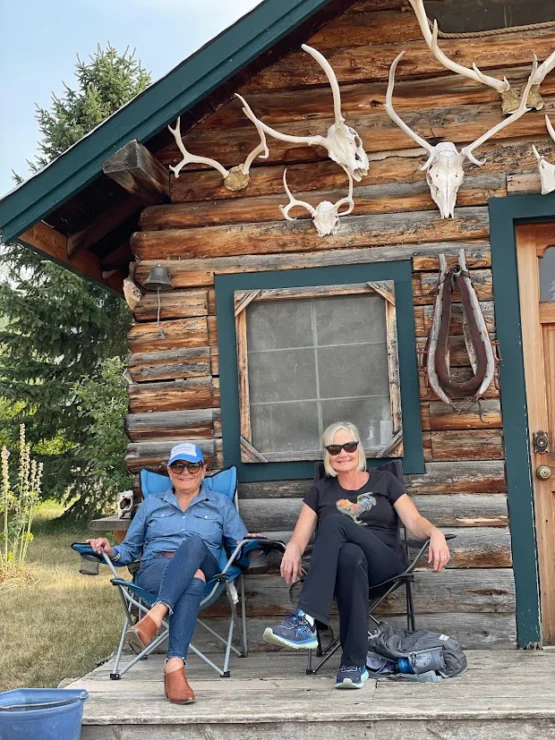 Two people sitting on folding chairs outside a log cabin with animal skulls mounted on the wall.