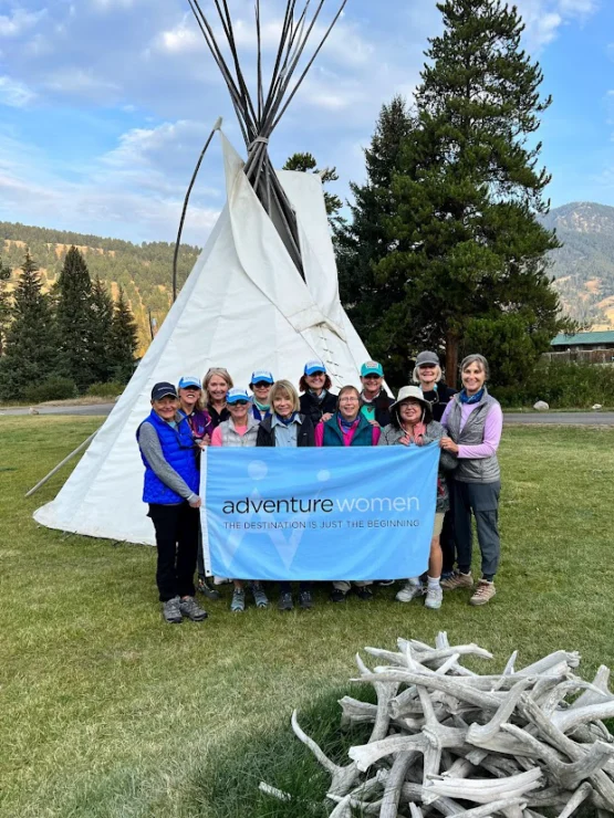 A group of people poses outdoors in front of a tipi, holding a banner that reads "Adventure Women: The Destination is Just the Beginning." Trees and mountains are visible in the background.