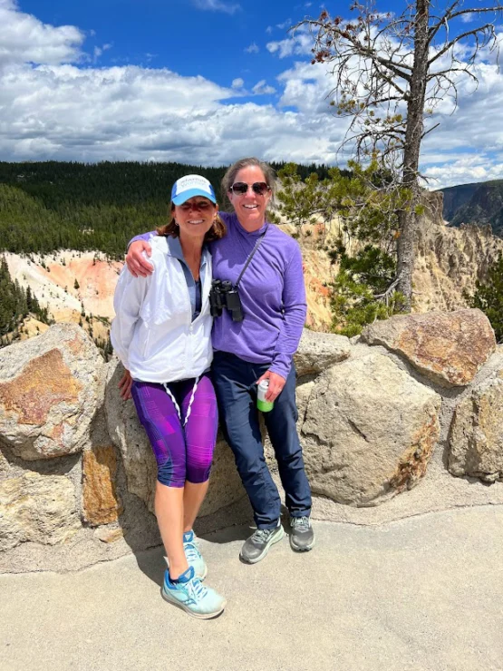 Two people in outdoor gear pose for a photo against a stone wall with a scenic canyon view in the background.