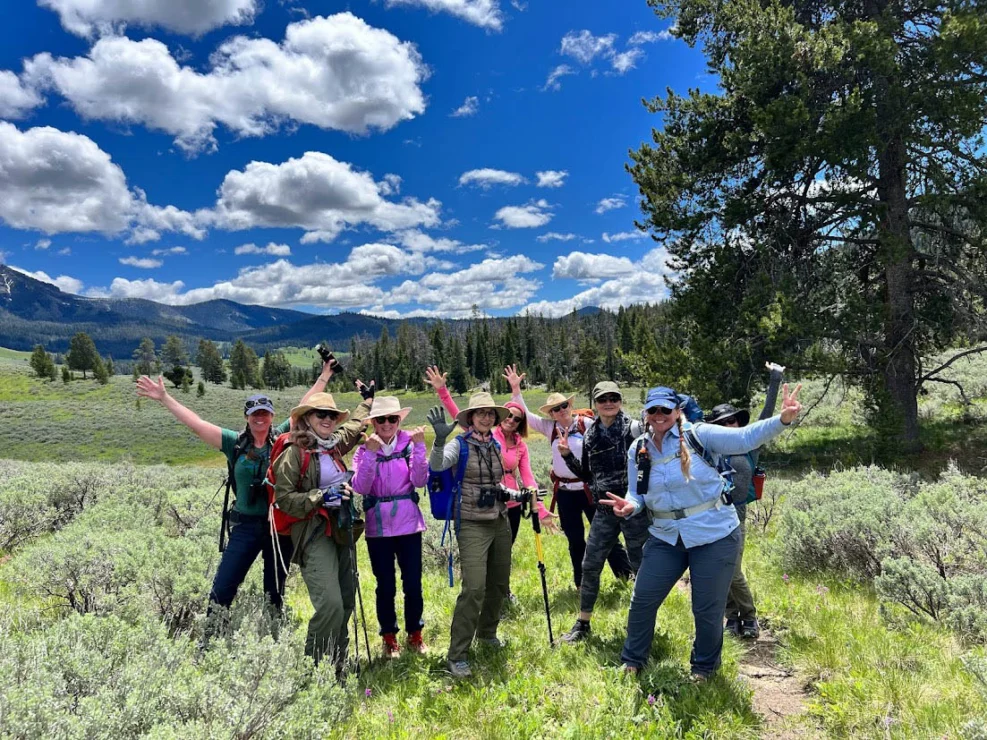 A group of hikers posing in a grassy field with trees and mountains in the background under a blue sky.