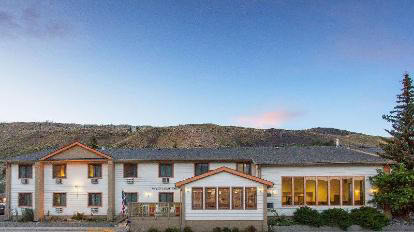 A two-story white building with a gabled roof stands against a backdrop of hills and a clear sky, with windows on both floors and a small entrance area at the front.