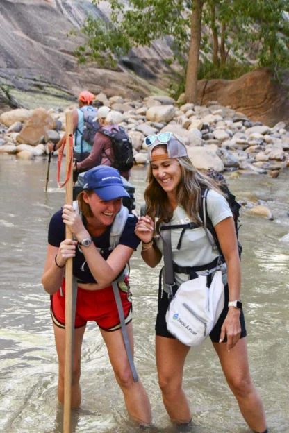 Two people smiling and wading through a rocky river with walking sticks. Others are visible in the background. They appear to be hiking in a natural setting with trees and large rocks.