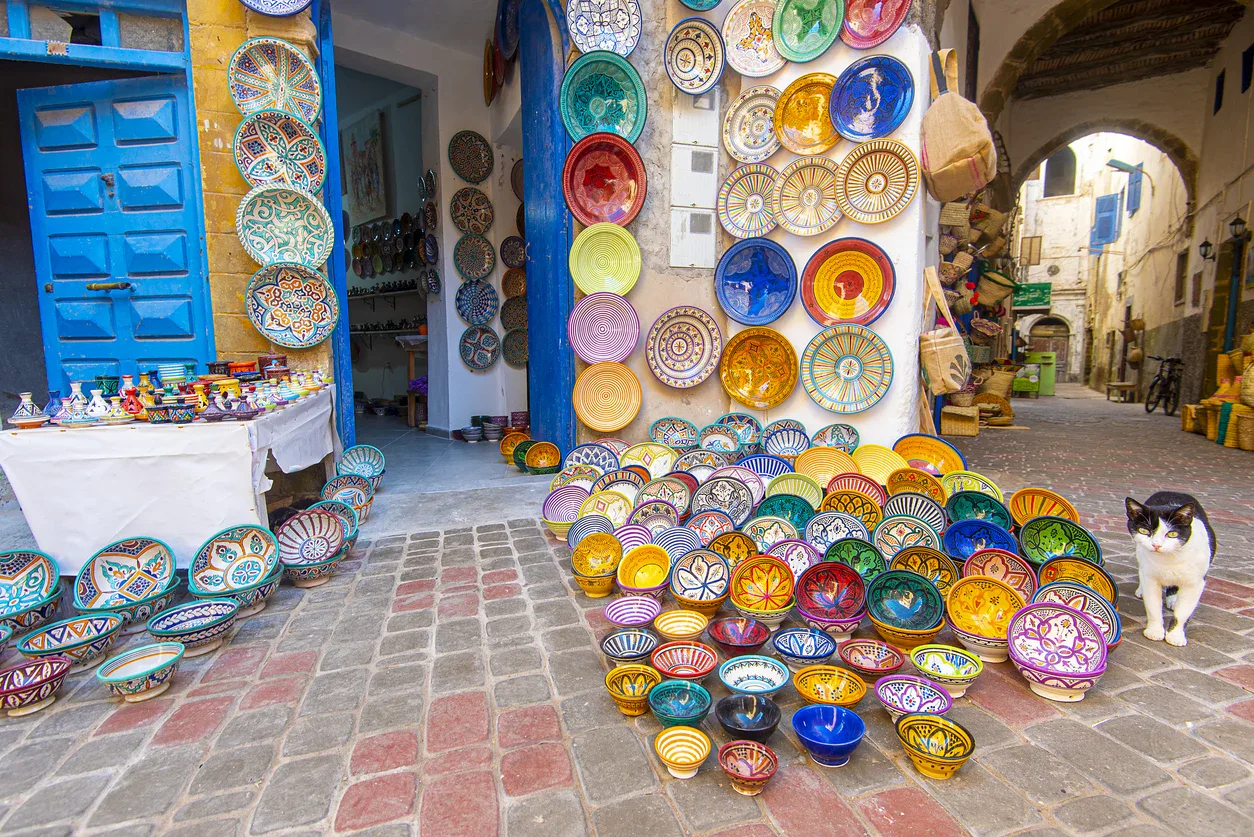 Colorful ceramic bowls and plates displayed outside a shop with a blue door on a cobblestone street; a black and white cat sits nearby.