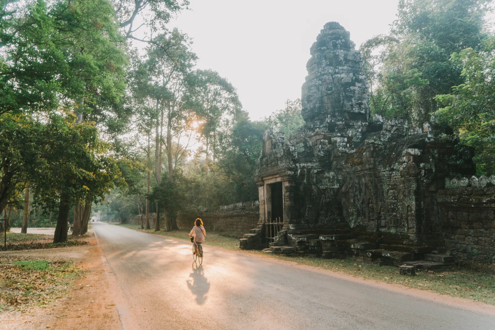 Person riding a bicycle on a road next to an ancient stone structure surrounded by trees.