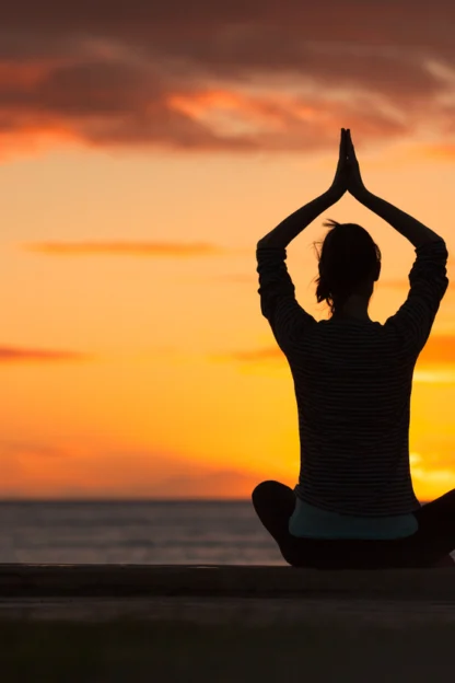 A person sits cross-legged in a yoga pose with hands raised above their head, silhouetted against an orange sunset by the water.
