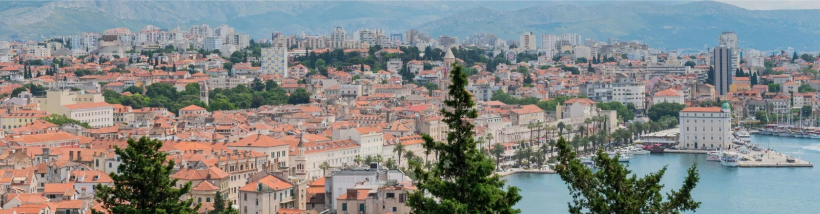 Panoramic view of a coastal city with red-roofed buildings, surrounded by green trees and hills in the background, with a body of water on the right.