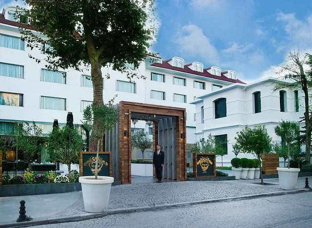 the entrance gate of a modern white hotel building with multiple windows and landscaped greenery.